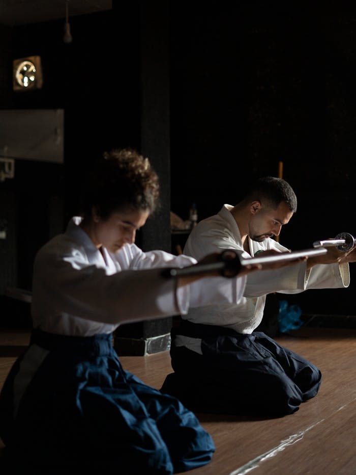 Two people in traditional martial arts attire practicing with swords indoors, focused and calm.