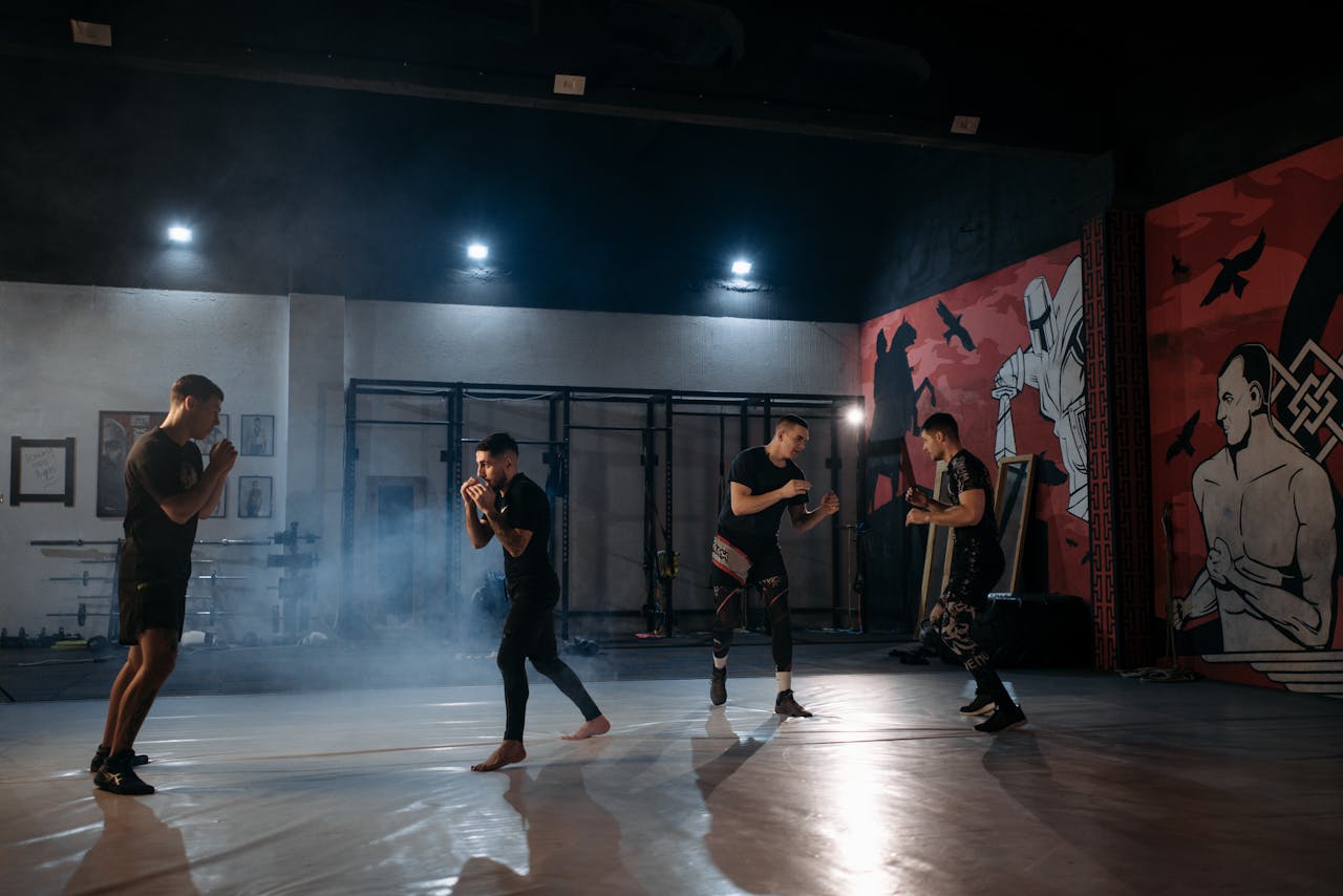 Four men sparring in a dimly lit gym with martial arts murals on the walls.