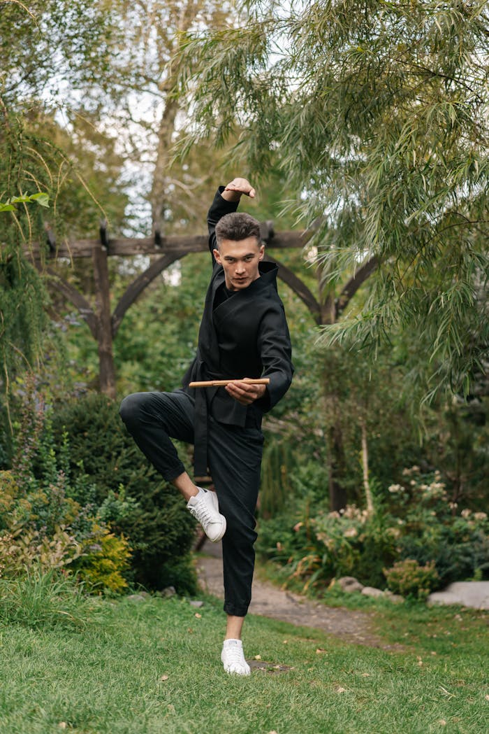about-us Martial artist performing a pose with a stick in a vibrant garden, showcasing strength and balance.